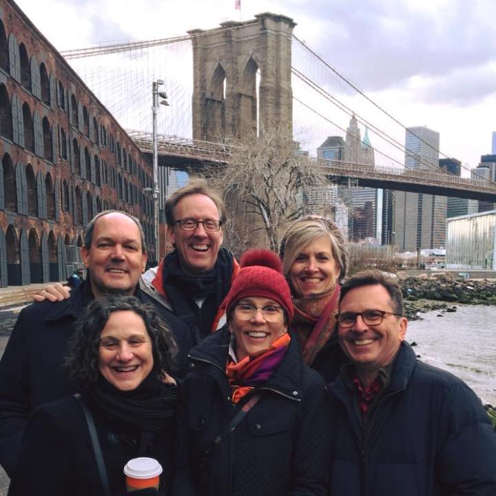 Group of six smiling adults posing together in front of the Brooklyn Bridge on a winter day during The Brooklyn Tour.